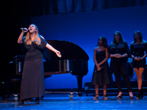 Female student singing on stage with backup singers and piano behind her